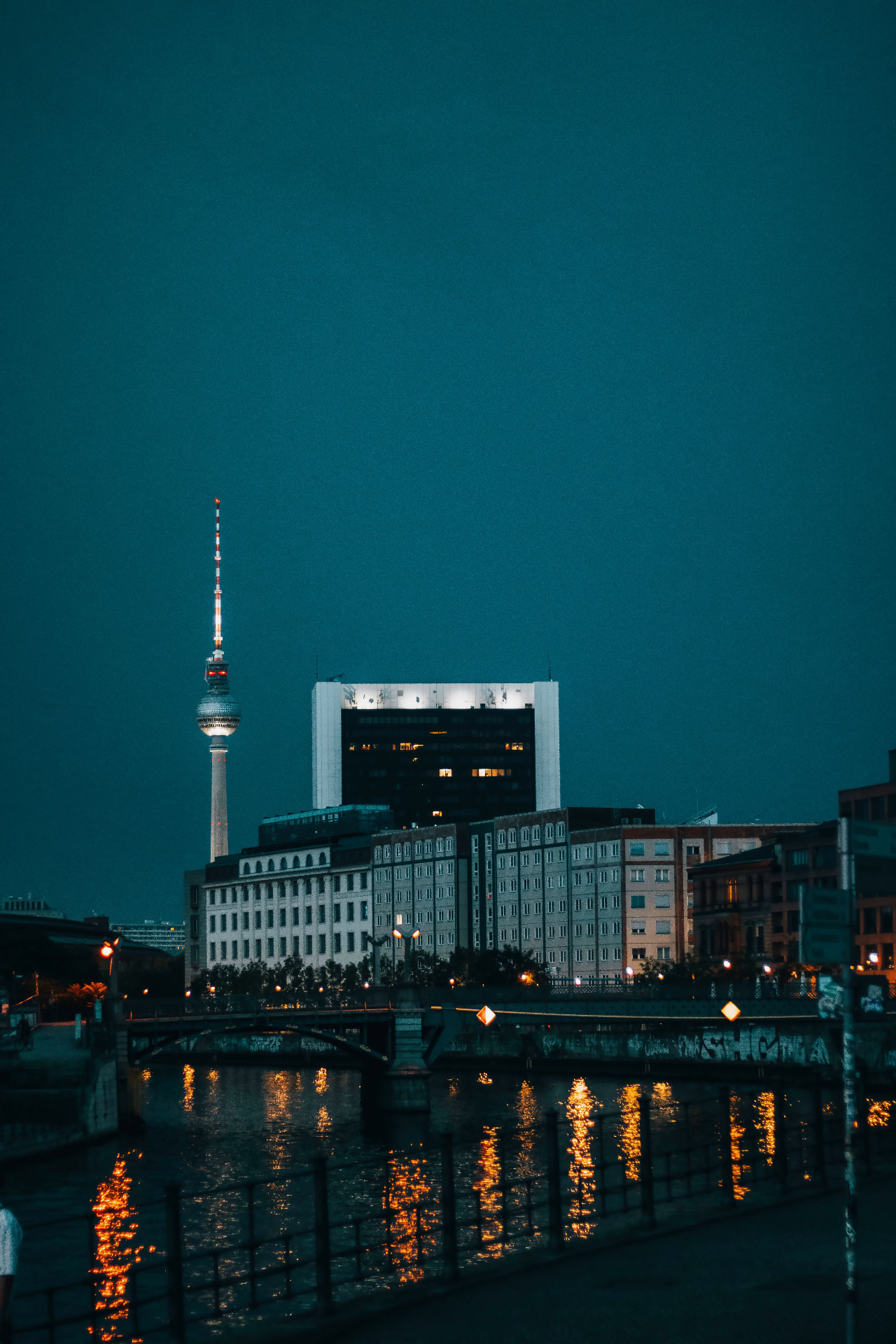 Berlin at blue hour, the illuminated TV Tower rising behind riverside buildings with golden light reflections on the dark water of the Spree