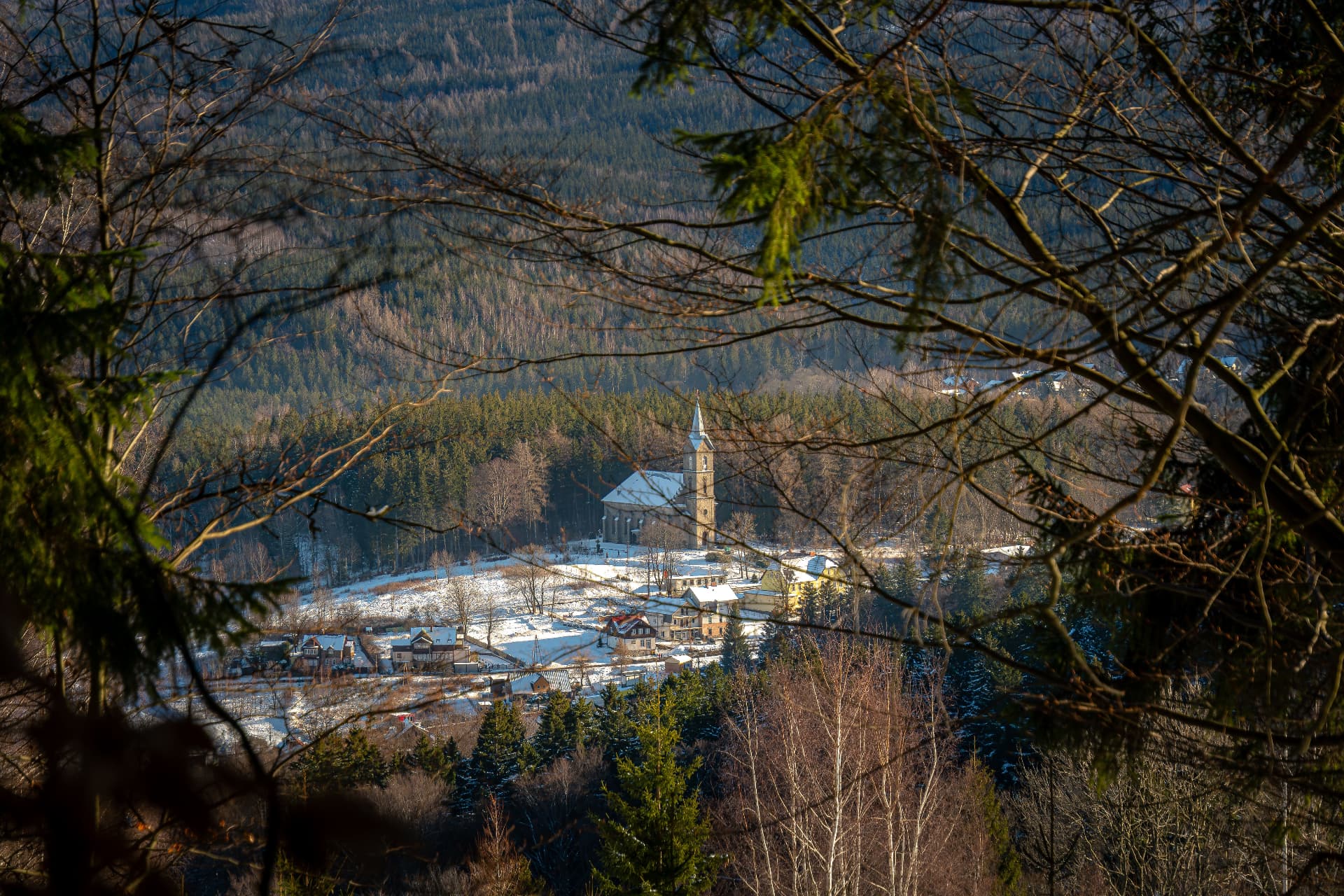 Snow-covered valley below Szklarska Poręba in the Karkonosze mountains of Poland, with a village church framed by pine branches