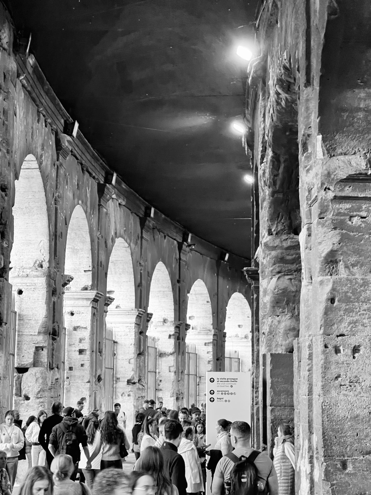 Black and white photograph of the Colosseum's upper arcade in Rome with visitors walking through