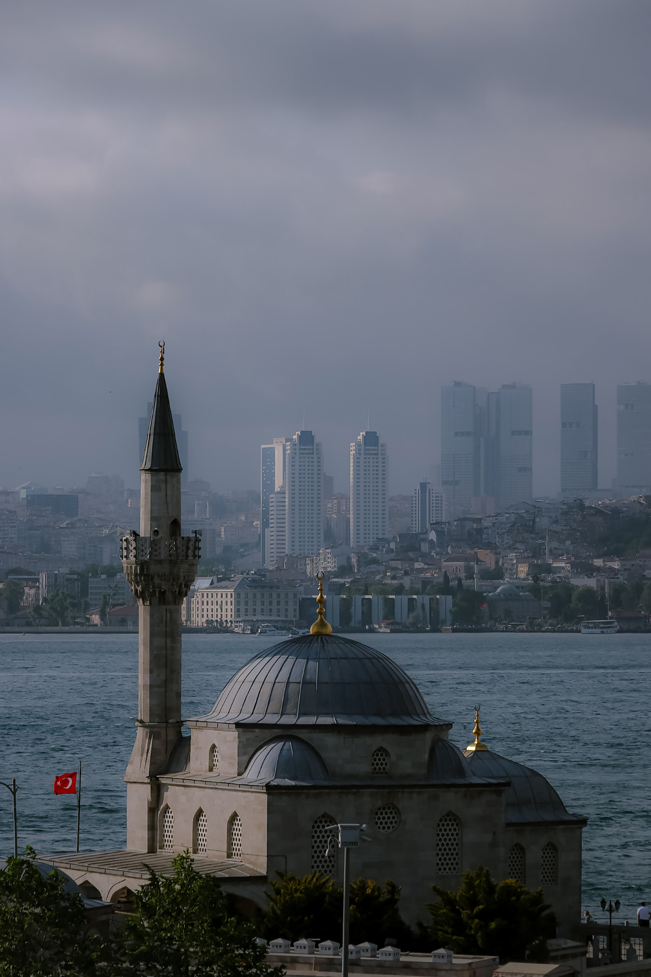 Şemsi Pasha Mosque on the Üsküdar waterfront in Istanbul with its dome and minaret against the modern skyscraper skyline across the Bosphorus under a hazy sky