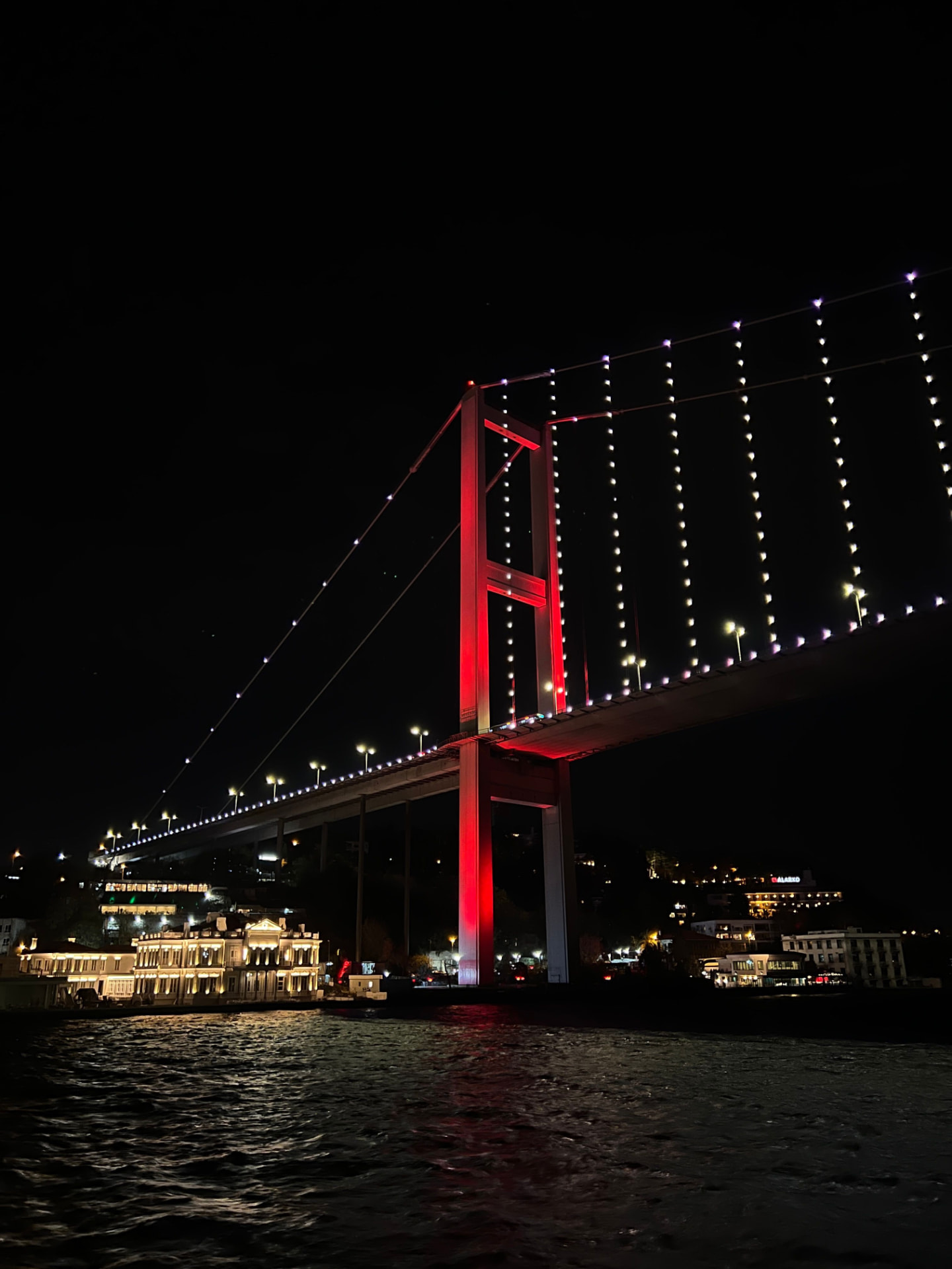 The 15 July Martyrs Bridge over the Bosphorus in Istanbul lit red at night, with an illuminated Ottoman-era waterside mansion below and reflections on the water