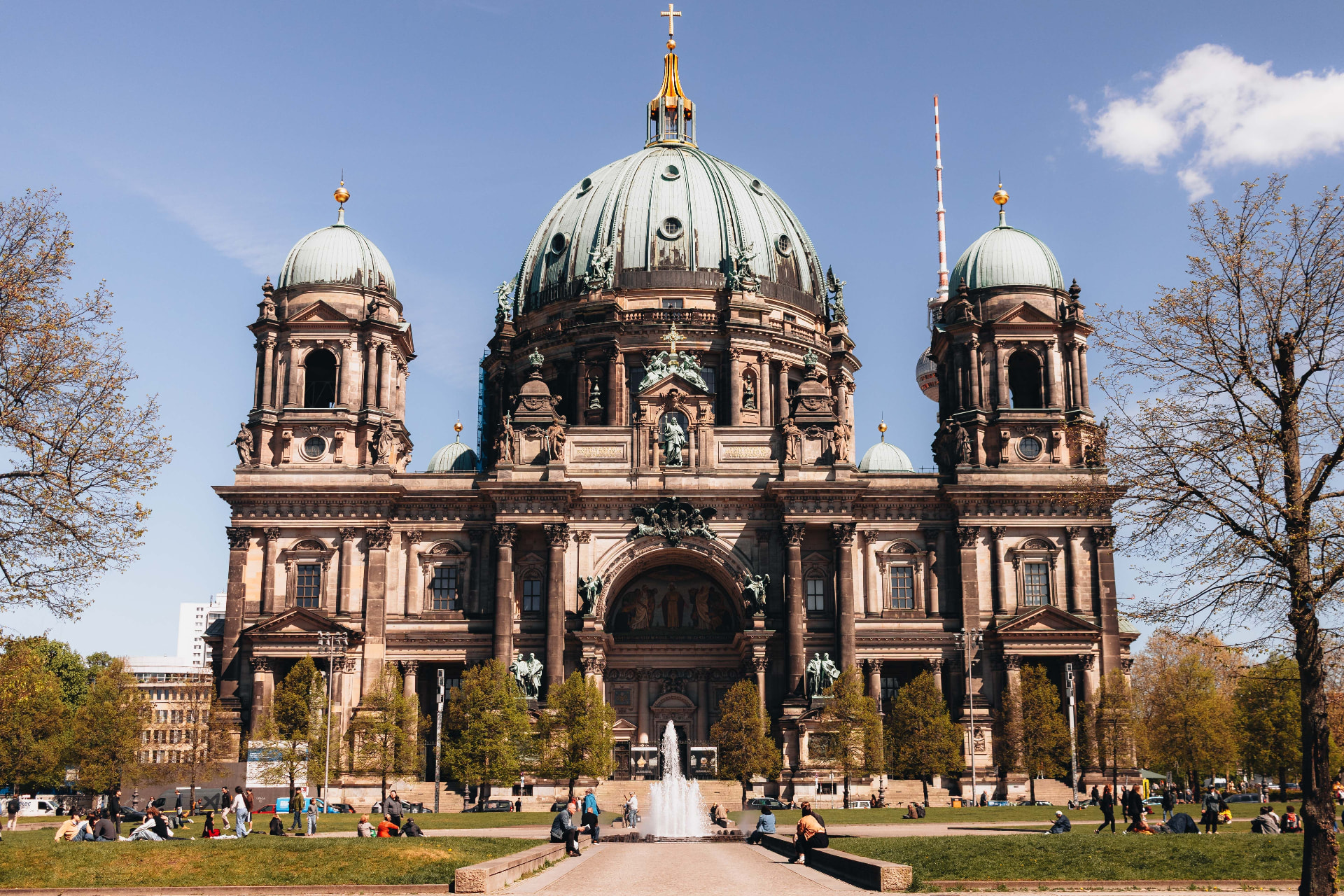 Berliner Dom cathedral in Berlin on a sunny spring day, its green copper domes against blue sky with a fountain and people on the Lustgarten lawn